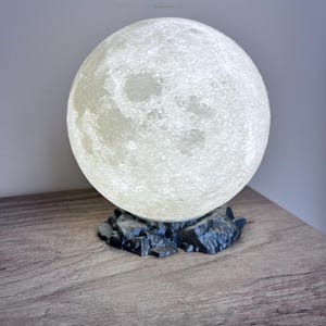 May include: A spherical, textured moon lamp with a bright, off-white glow. The lamp sits on a dark gray, rocky base. The lamp is on a wooden surface.