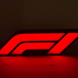 May include: A wall-mounted sign featuring the Formula 1 logo illuminated in a vibrant red light. The design consists of three slanted parallel lines on the left and a single diagonal bar on the right, creating the stylized F1 emblem against a dark background.