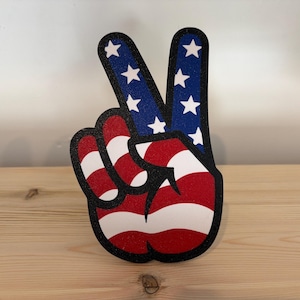 May include: A peace sign hand gesture in red, white, and blue, resembling the American flag. The fingers are outlined in black, with the index and middle fingers in blue with white stars. The palm features red and white stripes.