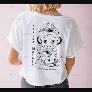May include: White t-shirt with a black graphic of Simba, Timon, and Pumbaa from The Lion King. The words "HAKUNA MATATA" are printed vertically on the left, and "IT MEANS NO WORRIES" is below the characters. A casual wear item.