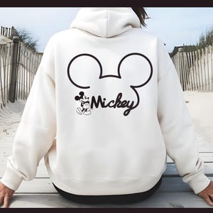 Könnte beinhalten: Weißer Hoodie mit einer schwarzen Mickey Mouse-Silhouette und dem Wort "Mickey" auf der Rückseite.