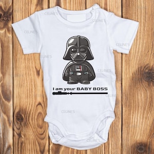 Könnte beinhalten: Weißer Baby-Body mit einem Cartoon-Motiv von Darth Vader. Die Illustration zeigt Vaders Helm und Rüstung. Darunter steht der Text "I am your BABY BOSS" mit einer Lichtschwert-Grafik. Der Body hat kurze Ärmel und einen Druckknopfverschluss.
