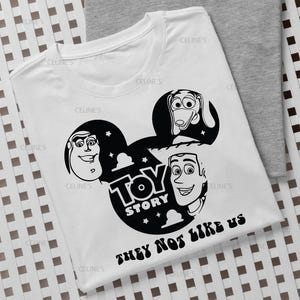 Puede incluir: Una camiseta blanca de manga corta con un gráfico negro de la silueta de la cabeza de Mickey Mouse que contiene ilustraciones de Buzz Lightyear, Slinky Dog y Woody. El texto dice Toy Story y They Not Like Us en una fuente negrita y ondulada.