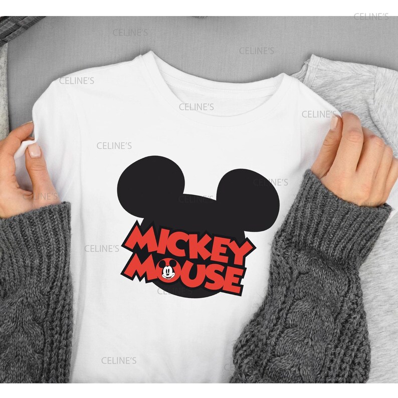 May include: White t-shirt with a black Mickey Mouse head design and the words "Mickey Mouse" in red. A small Mickey Mouse graphic is included. The shirt is held by hands wearing a grey sweater.
