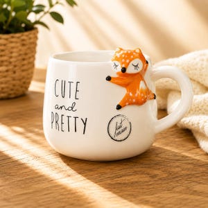 Peut inclure: Un mug en céramique blanche orné d'une figurine de renard orange et blanc en relief sur le côté. Le mug présente le texte noir "CUTE and PRETTY" ainsi qu'un tampon circulaire portant l'inscription "fait main" sur la face avant.