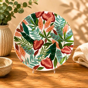 Peut inclure: Une assiette ronde en céramique ornée d'un motif botanique vibrant composé de feuilles tropicales superposées dans des tons vert foncé, bleu sarcelle clair et rose corail doux sur fond blanc, présentée sur un support en acrylique.