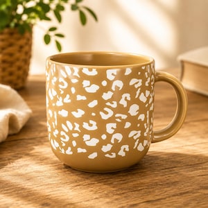 Peut inclure: Un mug en céramique jaune moutarde orné d'un motif répétitif en forme de taches de léopard blanches sur toute la surface extérieure. Ce mug présente un corps arrondi, une finition lisse et une anse incurvée sur le côté droit.