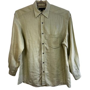 May include: A long-sleeved, button-down shirt in a muted olive green color. The shirt features a classic collar, a single chest pocket, and black buttons. The fabric appears to be a lightweight linen or similar material.