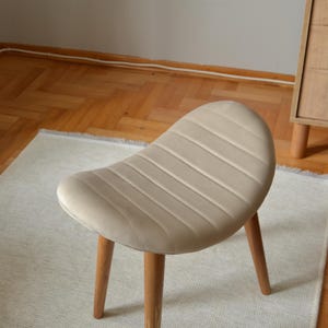 May include: A beige upholstered stool with a curved, padded seat and four wooden legs. The stool is sitting on a white rug.