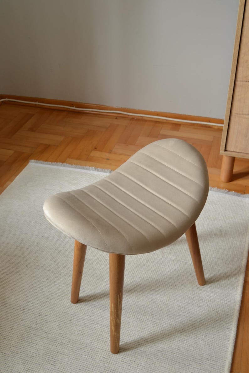 May include: A beige upholstered stool with a curved, padded seat and four wooden legs. The stool is sitting on a white rug.