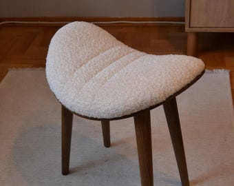 May include: A white upholstered stool with a curved seat and three wooden legs. The stool is on a light beige rug.