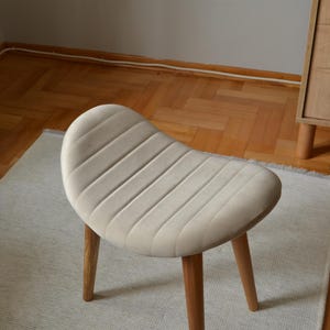 May include: A light beige upholstered stool with a curved, ribbed seat and four wooden legs.