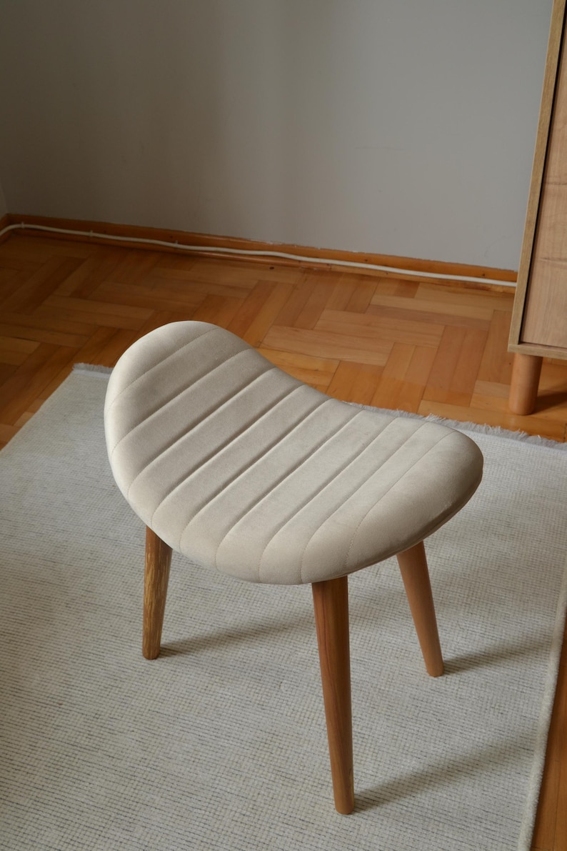 May include: A light beige upholstered stool with a curved, ribbed seat and four wooden legs.