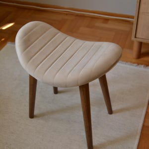 May include: A white upholstered stool with a curved seat and four wooden legs. The stool is sitting on a white rug.