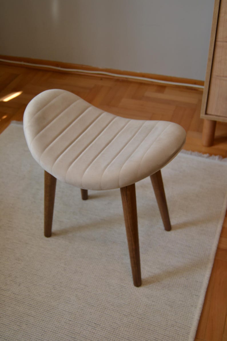 May include: A white upholstered stool with a curved seat and four wooden legs. The stool is sitting on a white rug.