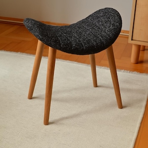 Puede incluir: Un taburete pequeño y moderno con un asiento texturizado gris oscuro y cuatro patas de madera marrón claro. El taburete está sobre una alfombra crema, con un suelo de madera y un armario en el fondo. El asiento tiene un diseño curvo y ergonómico.