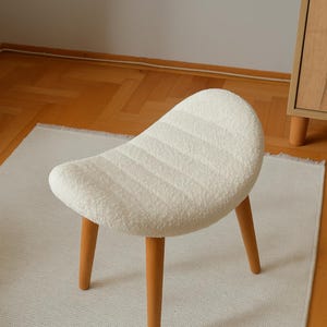 以下が含まれることがあります： 曲線的な座面と3本の木製の脚が付いた、小さな白い布張りのスツール。座面は、テクスチャ加工されたリブ模様です。脚は明るい茶色です。スツールは白いラグの上に置かれています。