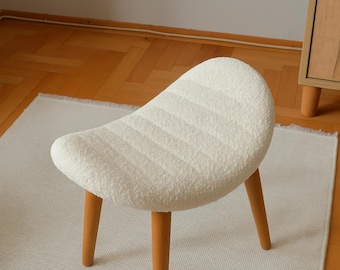 May include: A small, white, upholstered stool with a curved seat and three wooden legs. The seat has a textured, ribbed pattern. The legs are a light brown color. The stool is placed on a white rug.