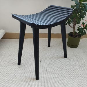 Könnte beinhalten: Ein schwarzer Hocker mit geriffelter Oberseite und vier sich verjüngenden Beinen. Der Sitz hat ein geschwungenes Design. Der Hocker besteht aus einem dunklen Material und steht auf einem hellen Teppich. Rechts steht eine Topfpflanze mit rosa Blüten.