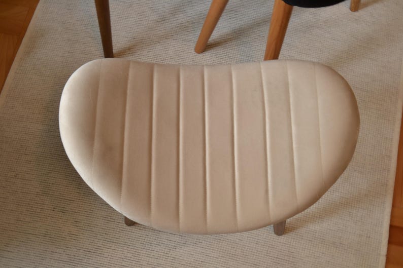 May include: A cream-coloured velvet chair seat with vertical stitching. The chair seat is oval shaped and has a smooth, soft texture.