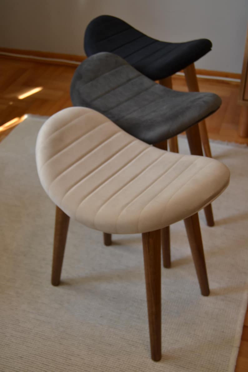 May include: Three upholstered stools with wooden legs. The stools are in different colours: beige, grey, and black. The stools are arranged in a staggered formation, with the beige stool at the front and the black stool at the back.
