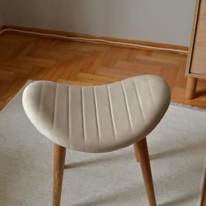 May include: A beige upholstered stool with a curved, ribbed seat and three wooden legs. The stool is sitting on a white rug.