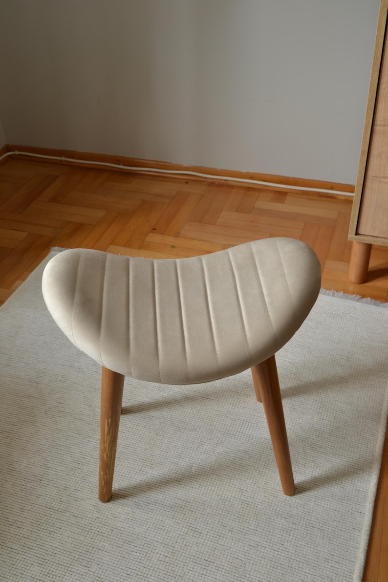 May include: A beige upholstered stool with a curved, ribbed seat and three wooden legs. The stool is sitting on a white rug.