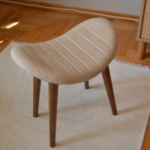 May include: A light beige upholstered stool with a curved seat and three wooden legs. The stool is sitting on a white rug.