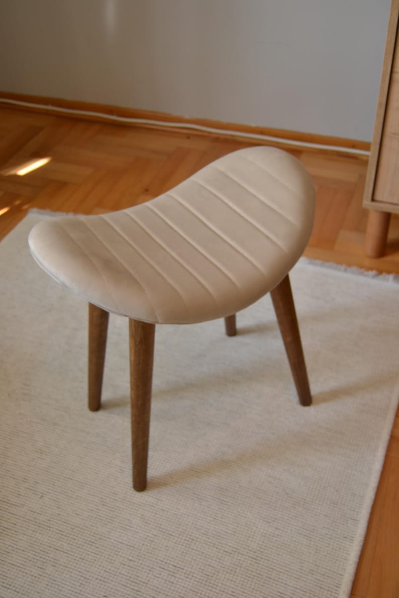 May include: A light beige upholstered stool with a curved seat and three wooden legs. The stool is sitting on a white rug.