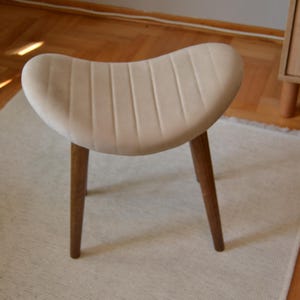 May include: A white upholstered stool with a curved, ribbed seat and three wooden legs.