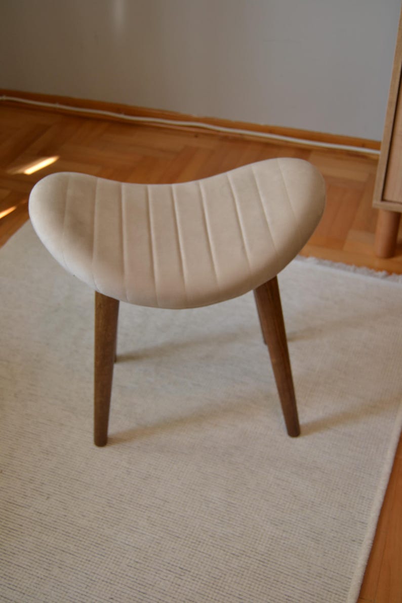 May include: A white upholstered stool with a curved, ribbed seat and three wooden legs.