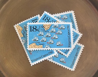 May include: A collection of vintage US postage stamps featuring a blue and white illustration of ships at sea. The stamps are from 1978 and depict the "Virginia Capes 1781" event. The stamps are arranged in a pile on a brass tray.