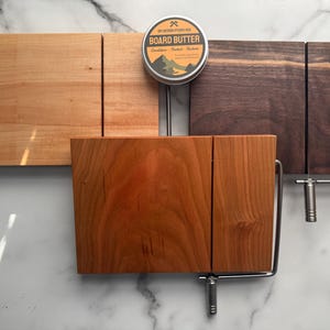 May include: Wooden cheese slicers in various wood tones, including light, medium, and dark brown. Each slicer has a stainless steel wire and handle. A small tin of "Board Butter" is centered above the slicers.