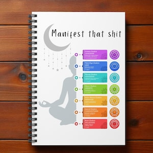 Op de afbeelding: Een wit spiraalgebonden notitieboek met een ontwerp met een mediterende figuur en chakrasymbolen. De tekst "Manifest that shit" staat boven een maan en sterren. Het notitieboek heeft een zwarte spiraalbinding.