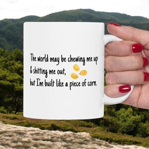 Puede incluir: Taza de cerámica blanca con una cita humorística sobre la resiliencia y el maíz. El texto dice: "El mundo puede masticarme y cagarme, pero estoy hecho como un trozo de maíz". Incluye un gráfico de maíz.