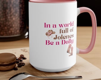 May include: White ceramic mug with a pink rim and handle. The mug has a pink and brown graphic with the text "In a world full of Jolenes Be a Dolly".