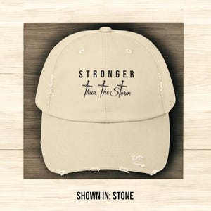 Puede incluir: Gorra de béisbol desgastada de color piedra con el texto "STRONGER Than The Storm" en negro. La gorra tiene una visera curva y un aspecto desgastado y vintage. La imagen también incluye el texto "SHOWN IN: STONE".