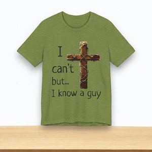 Könnte beinhalten: Grünes T-Shirt mit dem Text "I can't but... I know a guy." Das Shirt zeigt ein Design eines Holzkreuzes, das mit Stacheldraht umwickelt ist. Das T-Shirt wird auf einer Holzoberfläche präsentiert.