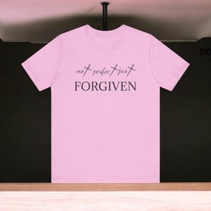 Puede incluir: Camiseta rosa claro con la frase "not perfect just FORGIVEN" en negro. Las palabras están separadas por símbolos de cruz. La camiseta tiene cuello redondo y mangas cortas. El fondo es oscuro.