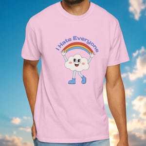 Op de afbeelding: Lichtroze t-shirt met een cartoonwolk die een regenboog vasthoudt met de tekst "I Hate Everyone". De wolk heeft een lachend gezicht en blauwe laarzen. Het shirt heeft een ronde hals.