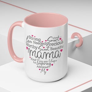 May include: White ceramic mug with a pink handle and pink interior. The mug has a design that says "mama" in a large font with the words "Strong, Love, Needed, Worthy, Sweet, Beautiful, Wise, Smart, Precious" surrounding it. The words "Kind, Fun, Inspiring, Admired, My" are below the word "mama" with a heart symbol after each word.