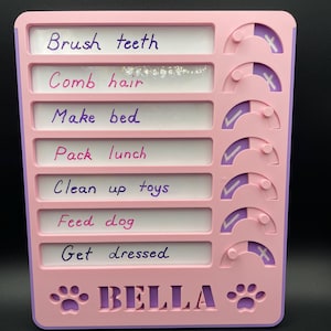 May include: A pink and purple dry erase chore chart with a paw print and the name "BELLA" at the bottom. The chart has seven rows with the following tasks: Brush teeth, Comb hair, Make bed, Pack lunch, Clean up toys, Feed dog, and Get dressed. Each task has a corresponding tick or cross to indicate completion.