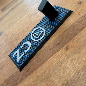 以下が含まれることがあります： ハニカム模様と「CZ」の文字とロゴが入った黒と青の長方形のオブジェクト。黒い長方形のブロックが上部に取り付けられています。オブジェクトは木製の表面にあります。