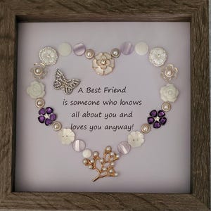 May include: Framed artwork featuring a heart-shaped arrangement of decorative buttons, pearls, and a butterfly. The text reads "A Best Friend is someone who knows all about you and loves you anyway!" The frame is a light brown color.