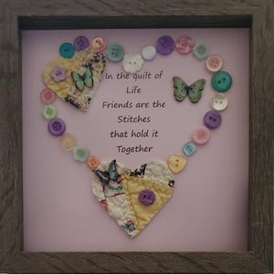 Könnte beinhalten: Gerahmtes Kunstwerk mit rosa Hintergrund und herzförmigem Design aus Knöpfen. Das Herz umgibt den Text "In the quilt of Life Friends are the Stitches that hold it Together". Zwei Stoffherzen und Schmetterlingsakzente vervollständigen das Design.