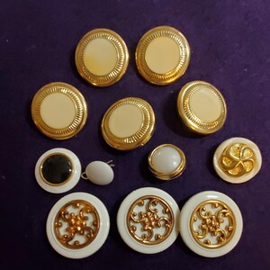 May include: A selection of vintage buttons in various sizes and designs. The buttons feature gold-toned metal rims and accents, with cream, black, and white centres. Some have ornate gold filigree patterns, while others are plain.