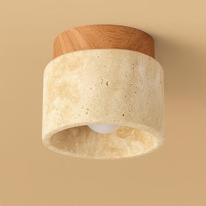 May include: A cylindrical ceiling light fixture with a light bulb. The fixture has a light beige, textured stone body and a wooden top. The light bulb is visible at the bottom of the fixture.