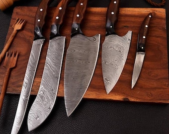 Puede incluir: Cinco cuchillos de cocina con mangos de madera y hojas con patrones se muestran en una tabla de cortar de madera. Los cuchillos están dispuestos en una fila, con el cuchillo más grande en la parte superior y el cuchillo más pequeño en la parte inferior.