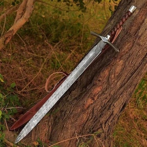 May include: A long, silver sword with a leather-wrapped handle. The sword has a decorative pattern on the blade and is resting on a tree trunk.