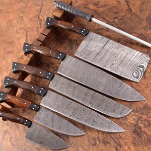 Può includere: Set di sette coltelli da cucina con manici in legno e lame in acciaio damascato. I coltelli sono disposti su un blocco di legno con un acciaio per affilare.